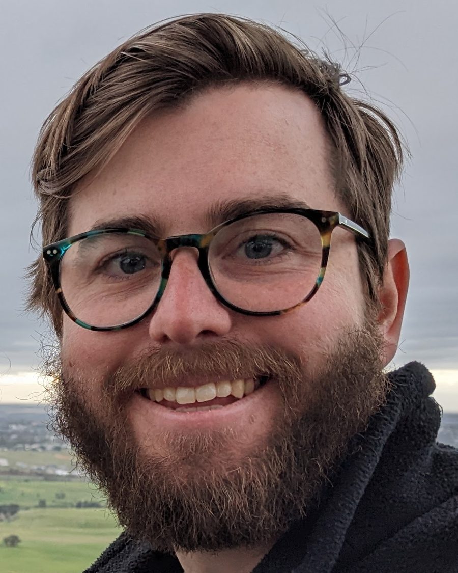Alex Preston smiling, wearing tortoiseshell glasses, on a hilltop
