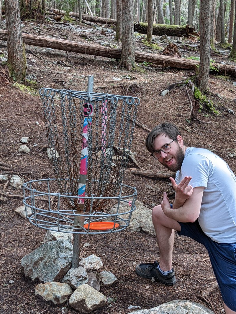 Alex crouching beside a disc golf basket in the forest, with an orange disc resting in it